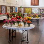A community hall filled with round tables displaying various floral arrangements and quilts hanging on the brick walls.